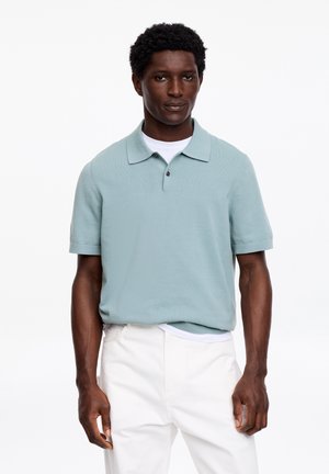 Man staat en draagt een lichtblauw poloshirt met korte mouwen over een wit T-shirt, gecombineerd met een witte broek, op een witte achtergrond.