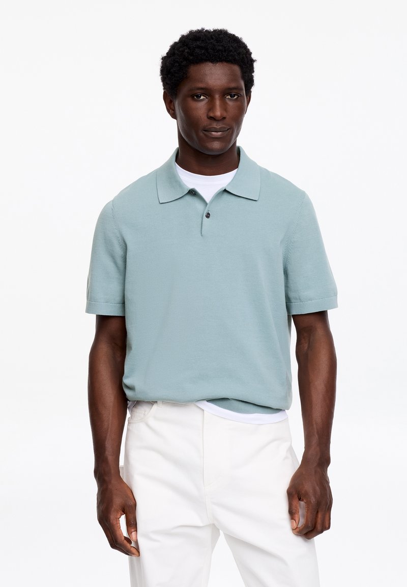Homme debout portant un polo à manches courtes bleu clair sur un t-shirt blanc avec un pantalon blanc sur un fond blanc.