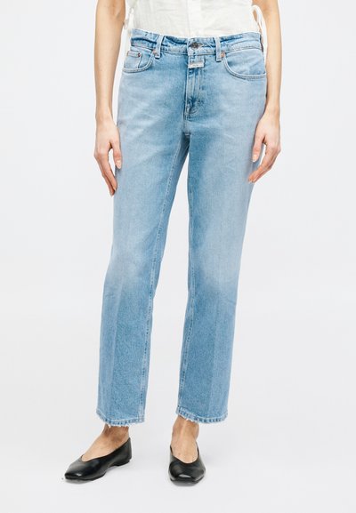Femme portant un jean bleu clair à jambes droites avec des ourlets bruts et des ballerines noires pointues, debout devant un fond blanc uni.