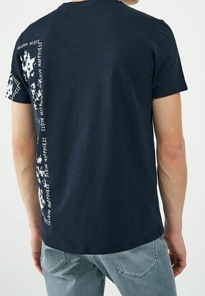 Uomo che indossa una t-shirt blu navy con testo bianco e grafiche sul retro sinistro e sulla manica, abbinata a jeans azzurri.