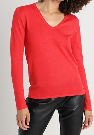 Femme portant un haut rouge vif à manches longues avec un col en V et un pantalon en cuir noir, debout les bras détendus le long du corps.