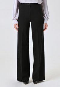 Schwarze weite Hosen aus glattem Stoff mit hoher Taille und einem dezenten vorderen Bundfalten. Kombiniert mit einer hellgrauen Bluse und spitzen schwarzen Absätzen.