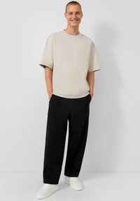 Beige Übergröße T-Shirt mit Rundhalsausschnitt, kombiniert mit lockeren schwarzen Hosen und weißen Sneakers, vor einem schlichten Hintergrund stehend.