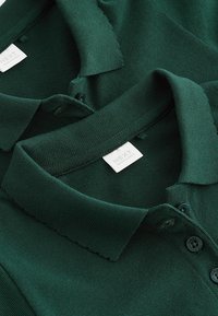 Tummanvihreät polo-paidat, joissa on teksturoitu kangas, klassinen kaulus, kolmipainikearvo ja etiketti, jossa lukee "NEXT."