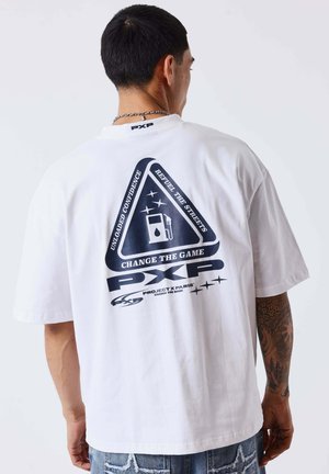 Man met kort zwart haar en een getatoeëerde arm, draagend een wit T-shirt met zwarte afbeelding en tekst op de rug, gecombineerd met een blauwe spijkerbroek.