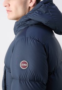 Tamno plava puffer jakna s visokim ovratnikom i vidljivim šavovima, koja sadrži okruglu oznaku Colmar na lijevom rukavu. Glatka tekstura.