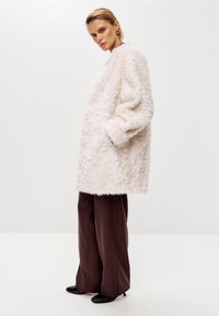 Faux fur frakke i lys pink med en struktureret, fluffy overflade. Løs pasform, sænkede skuldre og sidelommer. Parret med brune brede bukser.