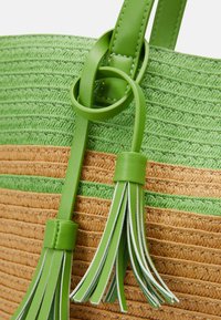 Bolsa tote de paja tejida en verde y marrón con acentos de cuero, que presenta un detalle anudado y borlas de cuero verde.