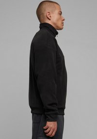 Svart fleece-pullover med høy krage, glidelås og ribbestrikkede ermer. Har en avslappet passform og glatt tekstur, kombinert med mørkegrå bukser.