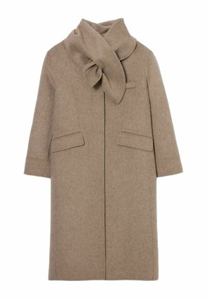 Donna che indossa un cappotto lungo beige con dettaglio sciarpa, camicia a quadri abbottonata, pantaloni larghi crema, scarpe marroni, che tiene una piccola borsa nera.