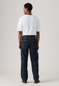 Vaqueros de denim en azul oscuro, con un ajuste relajado, dos bolsillos traseros con costuras en contraste y una etiqueta de la marca en el lado derecho. Combinados con una camiseta blanca.