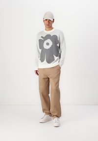 Hellgrauer Strickpullover mit großem dunkelgrauem Blumenmuster, beigen Hosen und weißen Sneakers. Weiche Textur mit gerippten Bündchen und Kragen.