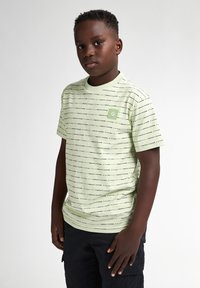 Camiseta verde claro con rayas negras horizontales, cuello redondo y un pequeño parche con un logo cuadrado en el lado izquierdo del pecho. Mangas cortas.