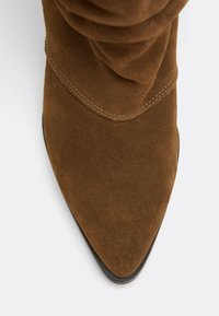 Botte en daim marron à bout pointu avec des coutures visibles autour de l’avant et une bordure de semelle noire sur fond blanc.