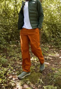 Vihreä vetoketjullinen toppatakki, valkoinen t-paita, oranssit cargo-housut ja harmaat vaelluskengät, esillä metsäisessä ympäristössä, jossa on vihreyttä.