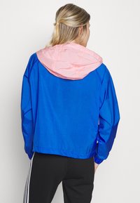 Chaqueta cortavientos azul con capucha rosa clara, que presenta un corte holgado, puños elásticos y una textura de tela suave y brillante.