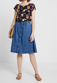 Mustat ylös Avon kukkakuviolla, sininen farkkuhame, jossa on napit, ja beige sandaalit. Hameessa on joustava vyötärö ja se on kellomainen.