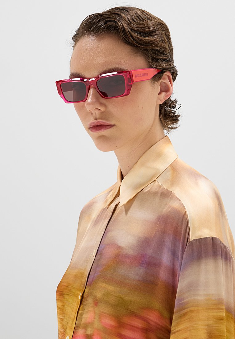 Lunettes de soleil en plastique rouge de forme rectangulaire avec des verres foncés ; "Just Cavalli" gravé sur la branche ; portées avec un haut en soie multicolore.