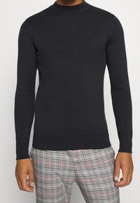 Mörkblå polotröja med långa ärmar, ribbade muddar och fåll. Materialet ser slätt ut med en slim fit. Buren över mönstrade rutiga byxor.