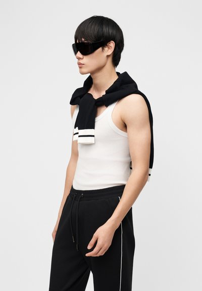 Νέος άνδρας που φοράει μαύρα γυαλιά ηλίου, λευκή φανέλα, μαύρη φόρμα με λευκές ρίγες και μαύρη ζακέτα περασμένη στους ώμους.