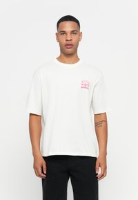 Jeune homme aux cheveux courts portant un T-shirt Adidas blanc avec un logo rose et un pantalon noir, se tenant devant un fond blanc uni.