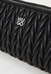 Pochette en cuir noir ornée d'un design matelassé avec des motifs torsadés. Logo argenté "HUGO" et fermeture zippée.