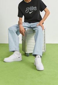 Svart t-shirt med vit Nike-logotyp, ljusblåjeans och vita Nike Air Force 1-sneakers. Sitter på en silverfärgad förvaringslåda med hjul.