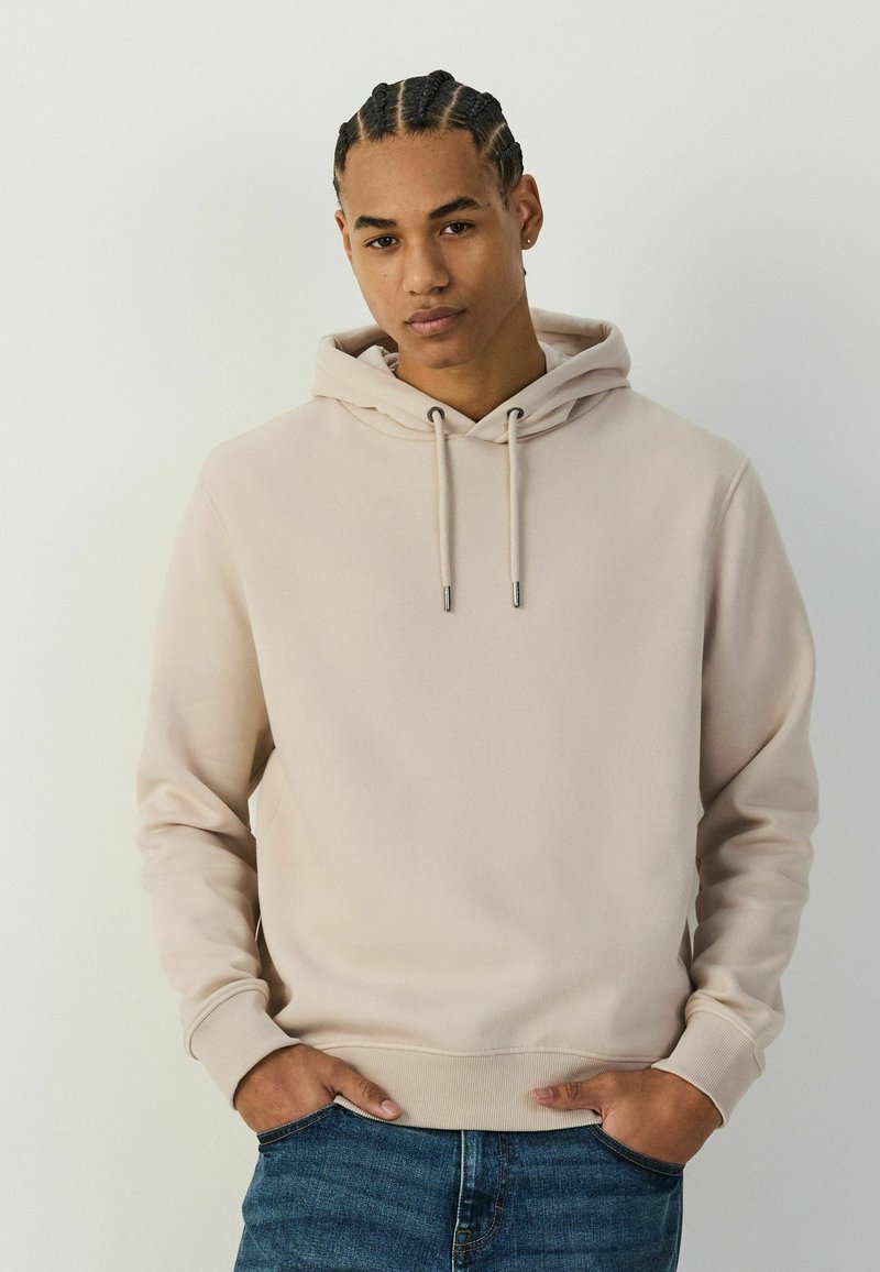 Jeune homme aux cheveux tressés portant un sweat à capuche beige et un jean bleu, debout les mains dans les poches devant un fond clair uni.