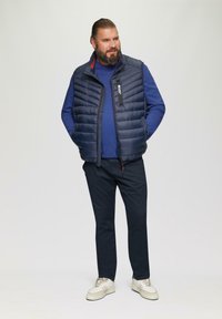 Marine gewatteerde vest over een blauwe lange mouwen shirt, gecombineerd met donkere broek en crèmekleurige sneakers. Heeft een rits en zakken voor opbergruimte.