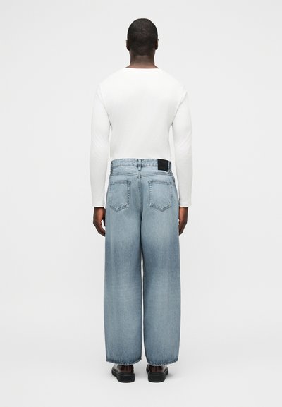 Άνδρας που φαίνεται από πίσω, φορά άσπρο μακρυμάνικο πουκάμισο, ανοιχτό μπλε τζιν παντελόνι με φαρδιά γραμμή και σκούρα παπούτσια, στέκεται μπροστά σε απλό φόντο.