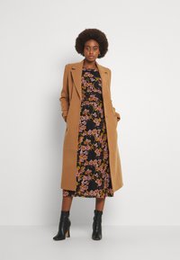 Cappotto di lana marrone sopra un vestito floreale nero con motivi gialli, rosa e verdi, abbinato a stivaletti neri aderenti alla caviglia.