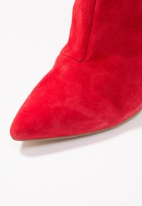 Stivaletto alla caviglia in suede rosso con punta affilata, che presenta una texture liscia e un design minimalista, evidenziando il colore vivace e la forma.