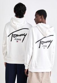 Sudaderas blancas con un gran logo negro de "Tommy" y el texto "JEANS" en la parte posterior, con puños y dobladillo acanalados.