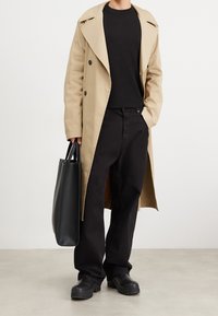 Beige trenčkot s širokými klopami, černý pletený svetr, černé kalhoty a robustní černé boty. Drž í černou kabelku. Hladká textura.