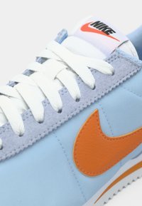 Plave platnene tenisice s detaljima od sivog antilopa, bijelim vezicama i narančastim Nike logom. Imaju teksturiranu potplat i podstavljeni jezik.