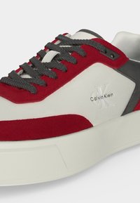 Λευκό, κόκκινο και γκρι αθλητικό παπούτσι Calvin Klein με σκουρόγκρι κορδόνια και ανάγλυφο λογότυπο στο πλάι πάνω από παχύ λευκό πάτο.