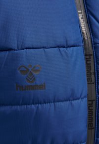 Giacca puffer blu con motivo a griglia, caratterizzata da un logo "hummel" nero e da un nastro di cerniera testurizzato lungo il bordo.