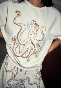 Biela mikina s jemnou výšivkou chobotnice v hnedej a modrej farbe, ku ktorej patria kraťasy s rovnakým dizajnovým motívom.