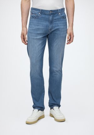 Mand iført lyseblå denimjeans og hvide sneakers, der står mod en ensfarvet hvid baggrund.