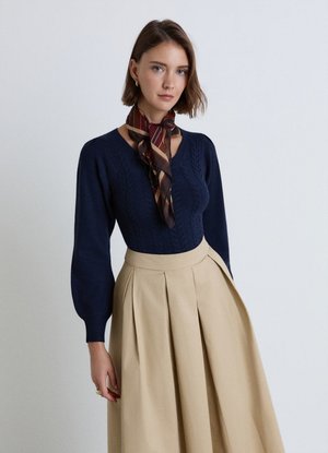 Donna che indossa un maglione a maniche lunghe blu scuro, una gonna beige plissettata e una sciarpa marrone a fantasia legata intorno al collo, in piedi davanti a uno sfondo semplice.