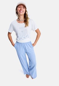 Camiseta blanca con diseño gráfico azul y acentos florales, combinada con pantalones anchos azul claro. La modelo lleva una gorra y un collar de conchas.