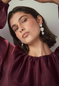 Frau mit kurzen dunklen Haaren, die große silberne herzförmige Ohrringe und eine durchsichtige burgunderfarbene Bluse trägt, posiert mit über dem Kopf erhobenen Armen.