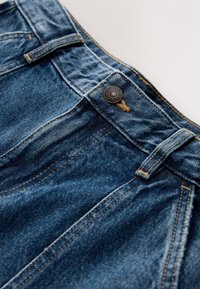 Blauwe denim jeans tailleband met metalen knoop met de tekst "NEX JEANS 1983" en zichtbare oranje stiksels op de riemlussen en naden.