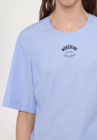 Light blue short-sleeve shirt with white vertical stripes and a round neckline. Features a black logo reading "Moschino Jeans" on the chest.