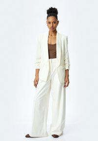 Blazer bianco su misura con ampi risvolti, abbinato a un top marrone aderente e pantaloni bianchi a gamba larga. Tessuto morbido, design minimalista.