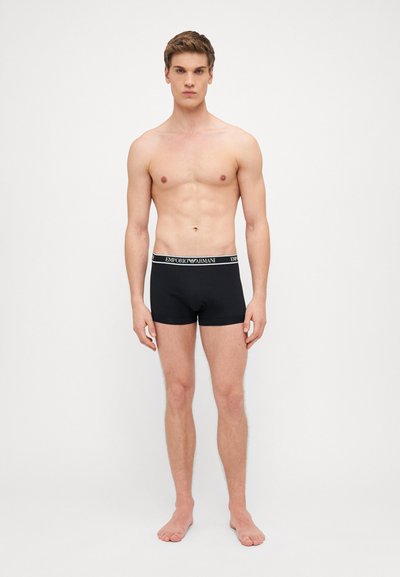 Černé boxerky s elastickým pasem a logem "Emporio Armani". Hladká tkanina a přiléhavý střih. Model stojí naboso.