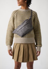Grå lærcrossbody-veske med teksturert overflate, med en frontlomme med glidelås og justerbar stropp, brukt over en stripete strikkegenser.