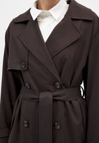 Mörkbrun trenchcoat med dubbelknäppt design, stor krage och ett knytbart midjebälte. Har dekorativa knappar och texturerad tyg.