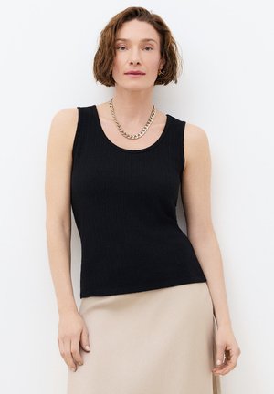 Nainen, jolla on lyhyet laineikkaat hiukset, musta hihaton yläosa, beige hame ja kultainen ketjukoru, seisoo valkoista seinää vasten.