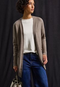 Beige stickad cardigan med ribbade manschetter, bärs över en vit skjorta och blå jeans. Modellen håller en ljusgrå handväska.
